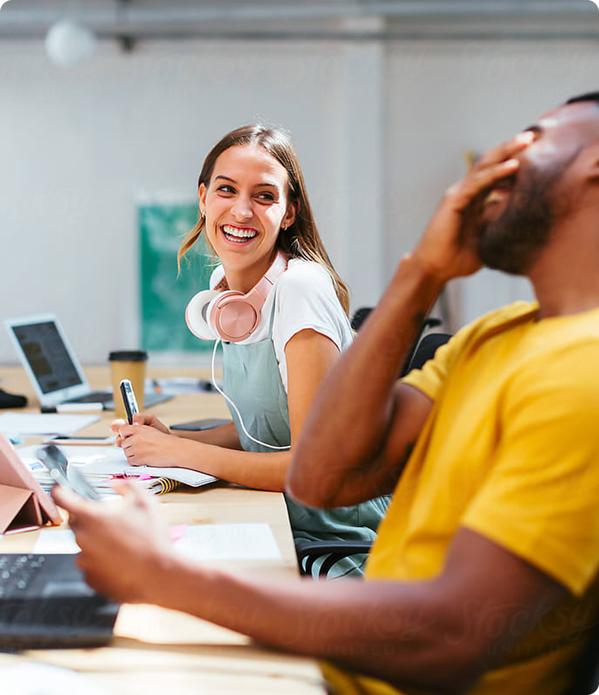 Two young adults laughing and working at a shared desk, with laptops, notebooks, and headphones, suggesting a fun and collaborative study or work session.