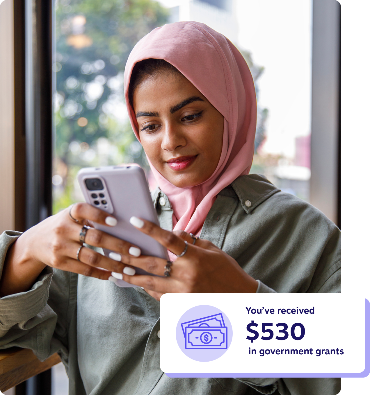 Young woman wearing a pink hijab looking at her phone by a café window, with a notification showing she received $530 in government grants.
