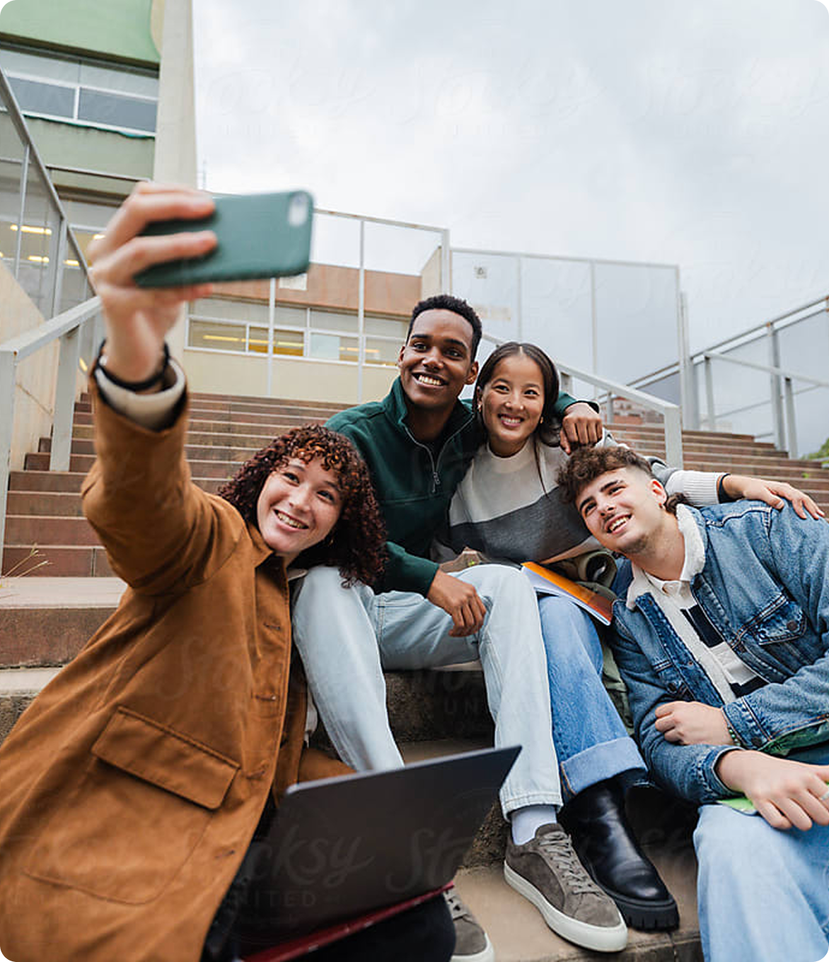 Group of four diverse university students sitting on outdoor steps, smiling and taking a selfie together with a phone and laptop nearby.