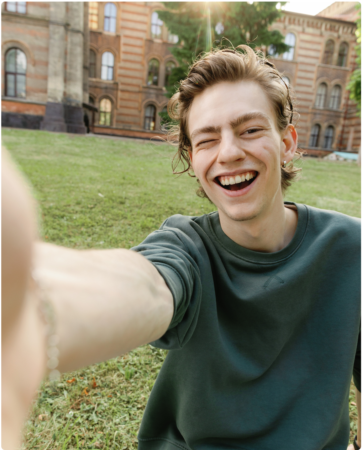 Trendy guy with cheery grin raising his arm, winking at camera and photographing himself in sunny weather across from educational establishment on its green area.