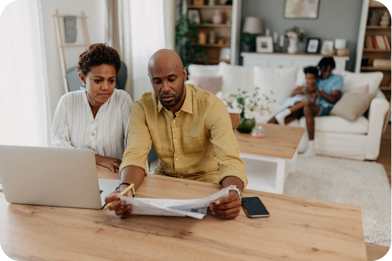 Mother and father reviewing financial documents on a laptop and reviewing papers while their children play in the background