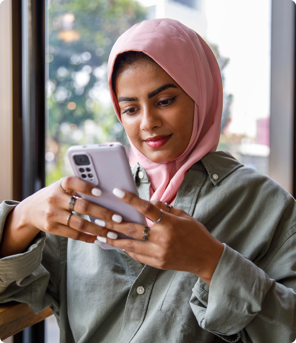 Young woman wearing a pink hijab looking at her phone by a café window.