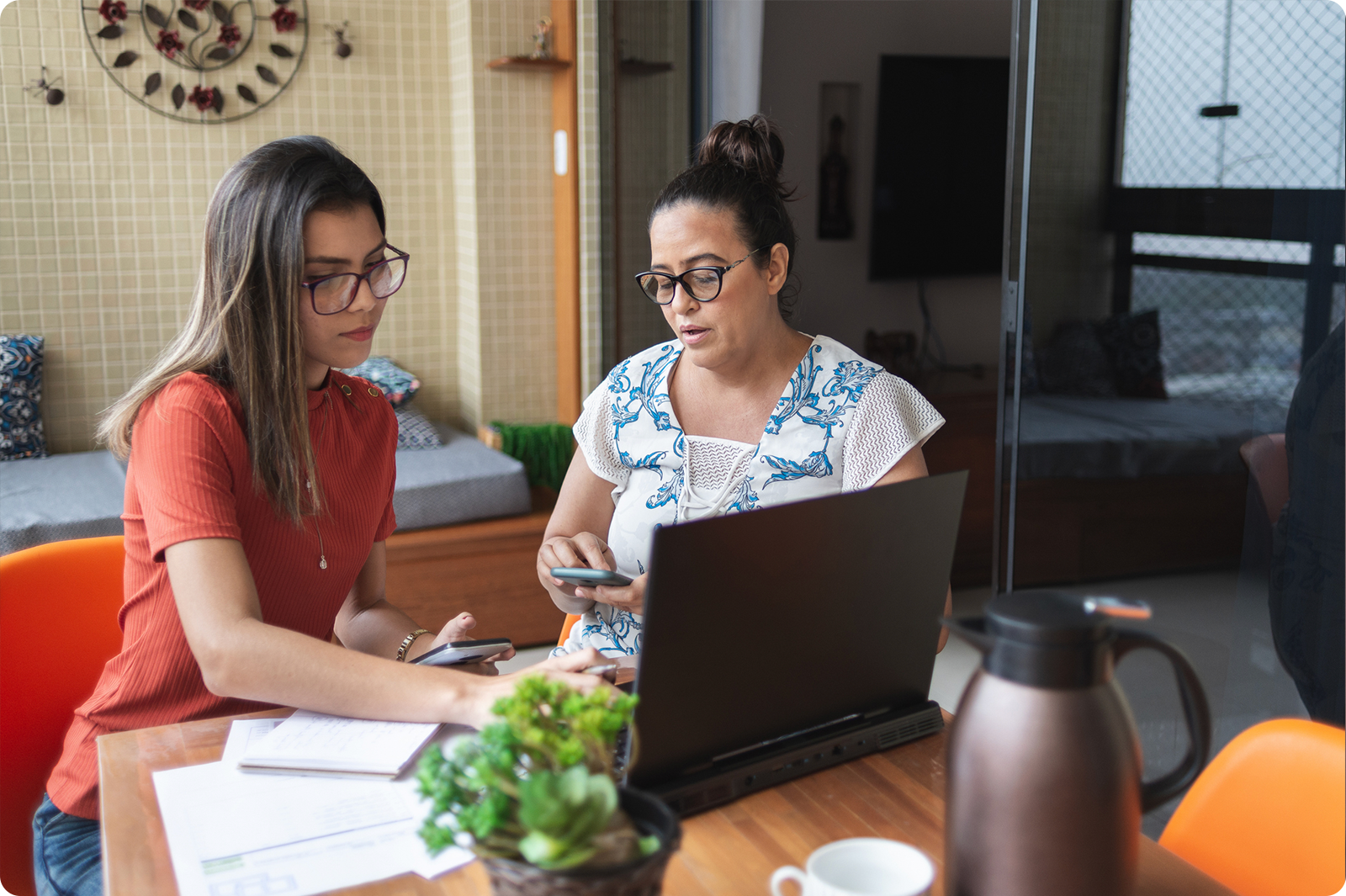 Mother and daughter working together at a wooden dining table with a laptop, both wearing glasses. One woman in a coral sweater holds a phone while the other in a white and blue patterned top looks at the laptop screen. A modern kitchen is visible in the background.