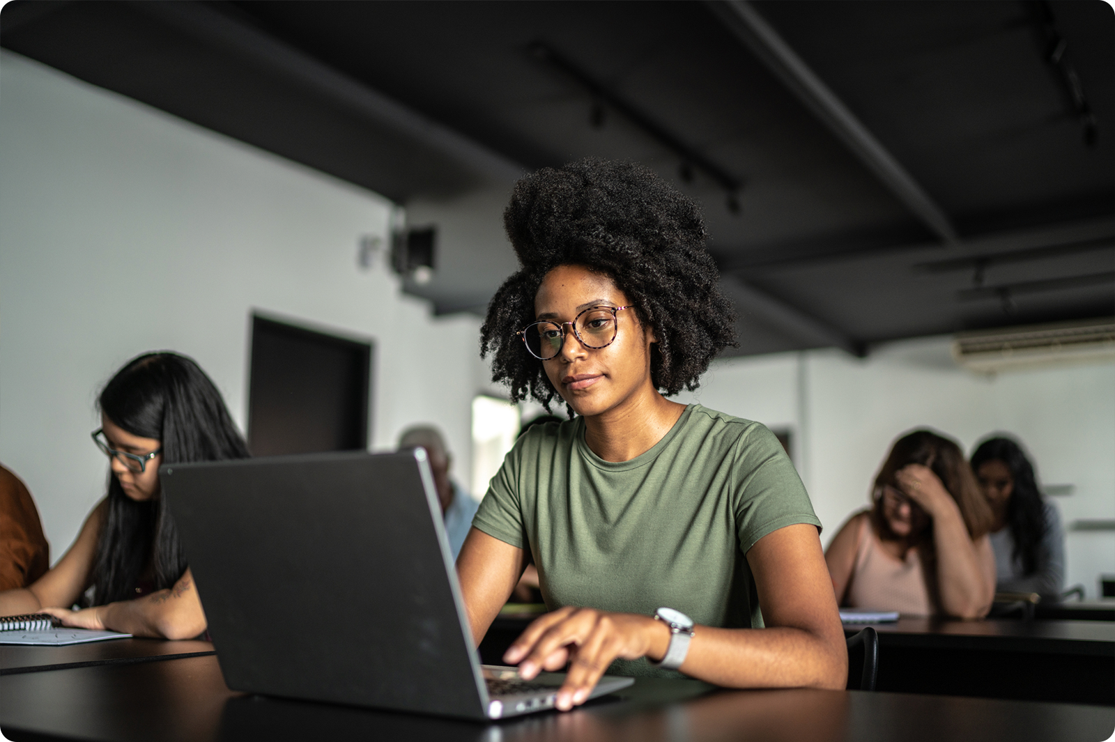 A Black woman with natural curly hair and glasses sits at a laptop in a modern office or coworking space. She's wearing a green t-shirt and appears focused on her work. In the background, other people are also working at laptops in the bright, contemporary workspace with exposed ceiling beams.