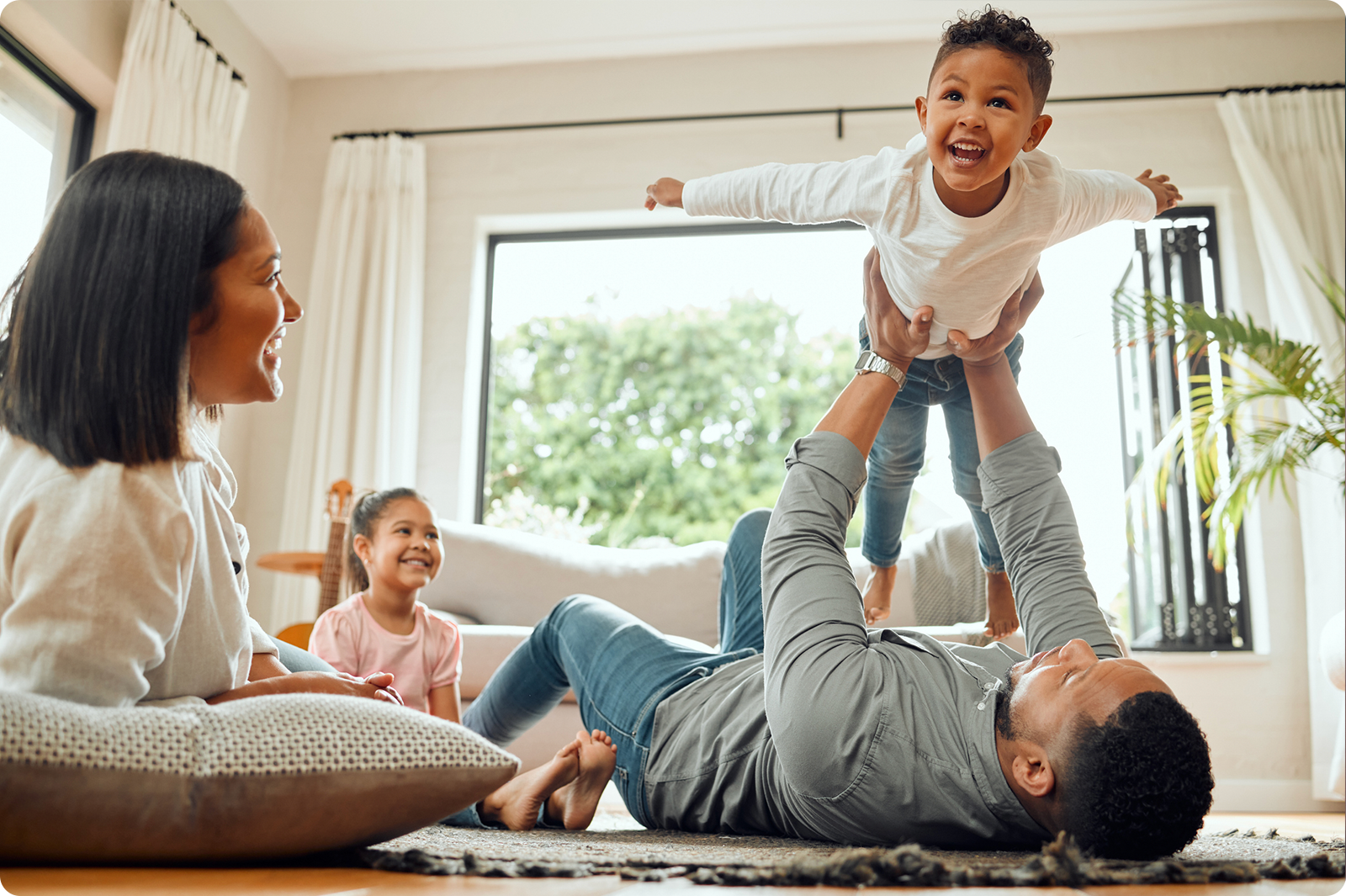 A joyful family scene with a father lying on the floor holding up his laughing toddler in the air while a mother and young daughter watch and smile from a nearby couch in a bright, modern living room.