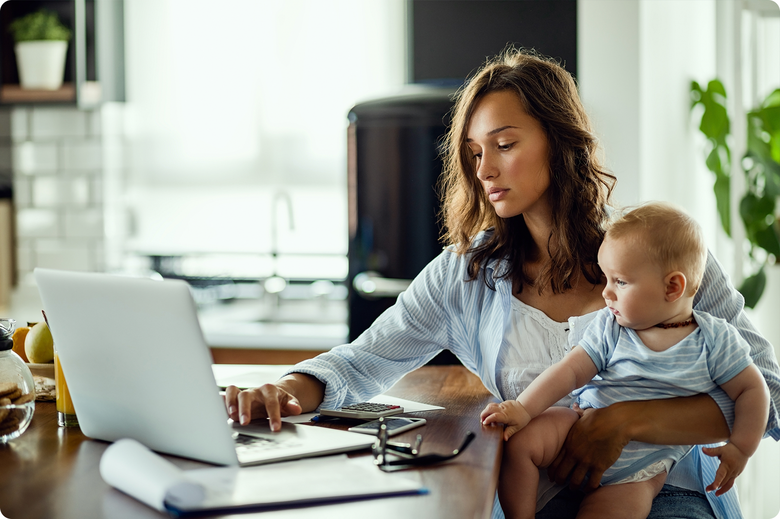 A mother with wavy brown hair working on a laptop while holding her baby on her lap. She wears a light blue striped shirt and sits at a wooden table in a bright, modern kitchen with plants and shelving visible in the background.