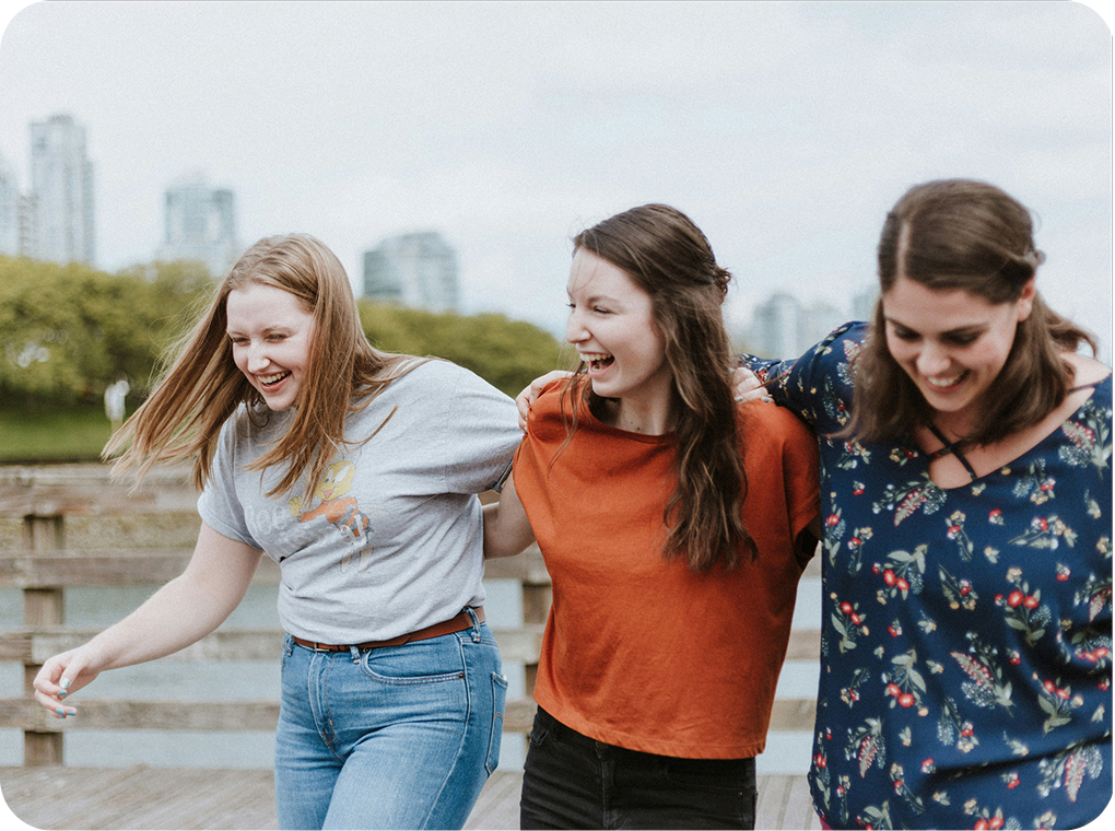 Three women joyfully laughing together on a bridge, enjoying a sunny day and each other's company