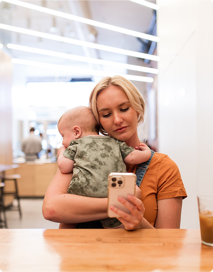 A woman cradles a baby in one arm while using a cell phone with the other hand, smiling gently.