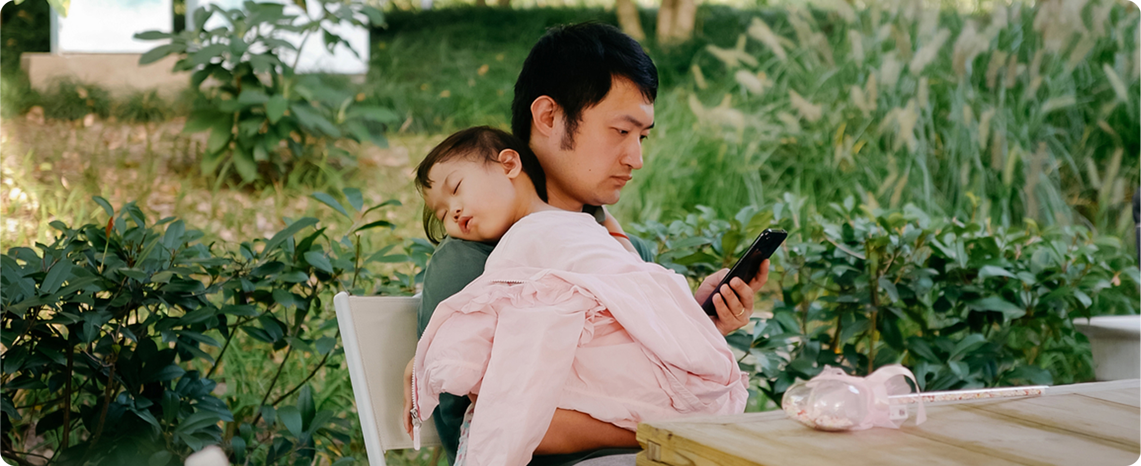 A man sitting on a bench, gently holding a baby in his arms, both smiling and enjoying a sunny day together.