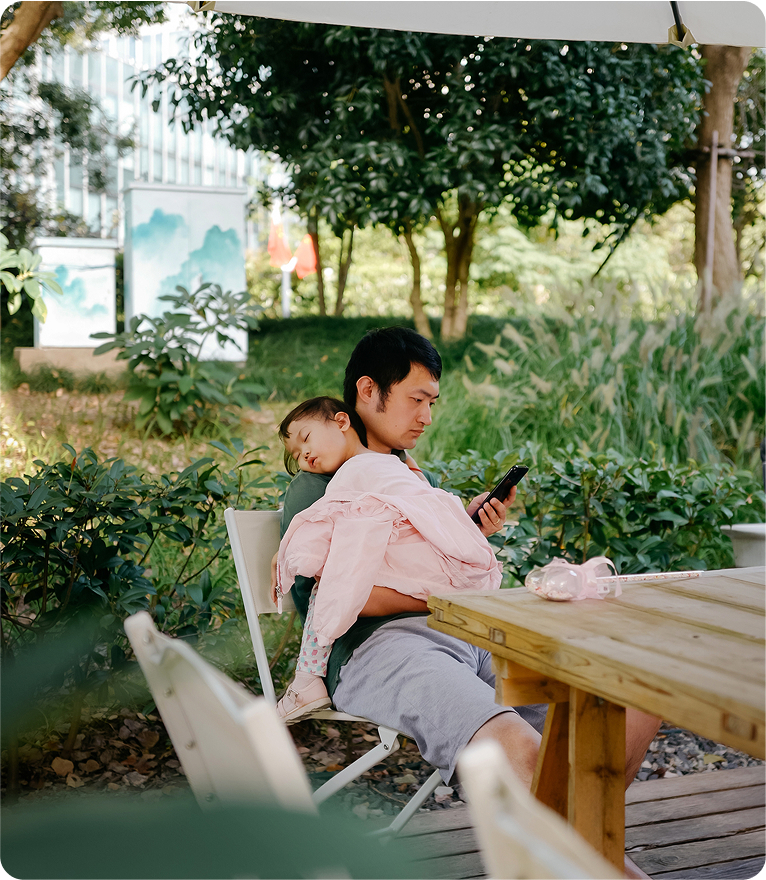 A man sitting on a bench, gently holding a baby in his arms, both smiling and enjoying a sunny day together.