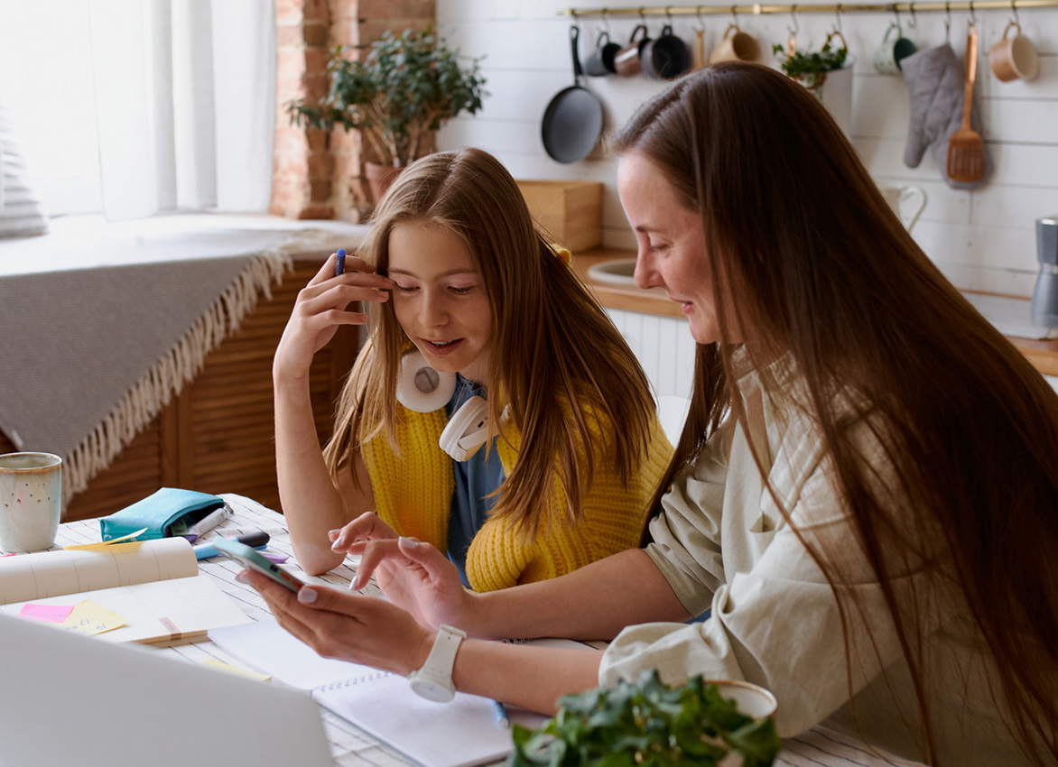 Mom and daughter sit at a table, engaged in conversation with a laptop and a phone in front of them.