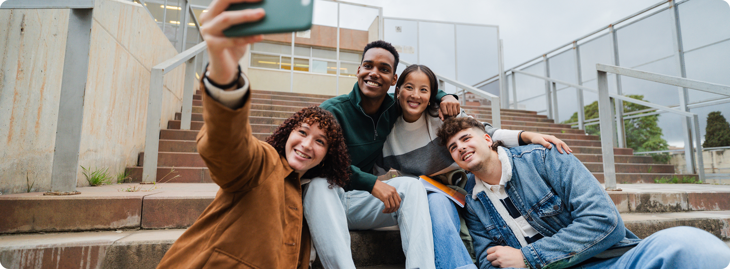 Group of four diverse university students sitting on outdoor steps, smiling and taking a selfie together with a phone and laptop nearby.